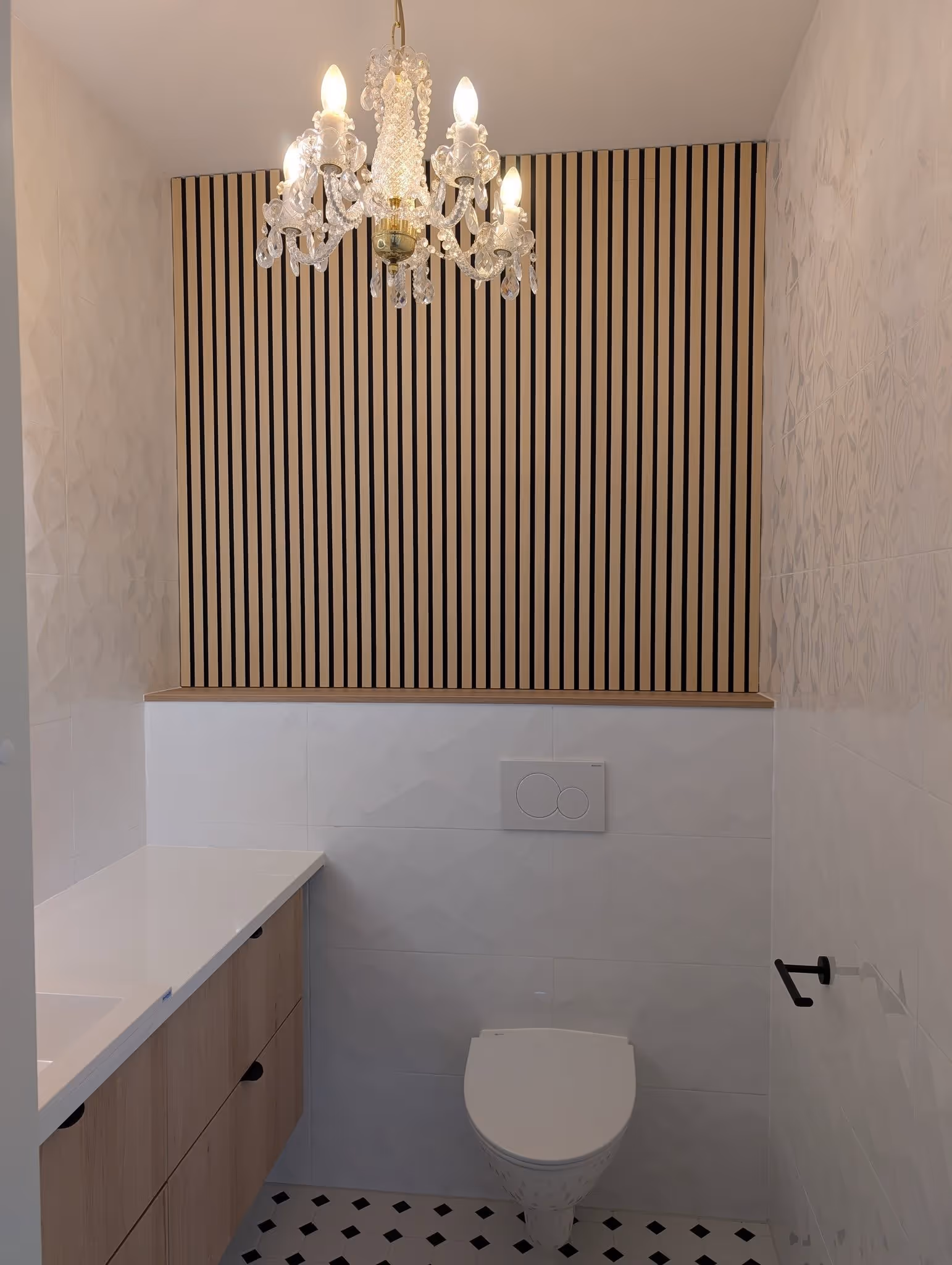 Kompakti kylpyhuone, jossa valkoinen seinä-WC, kristallikruunu ja vaaleat puukaapit.