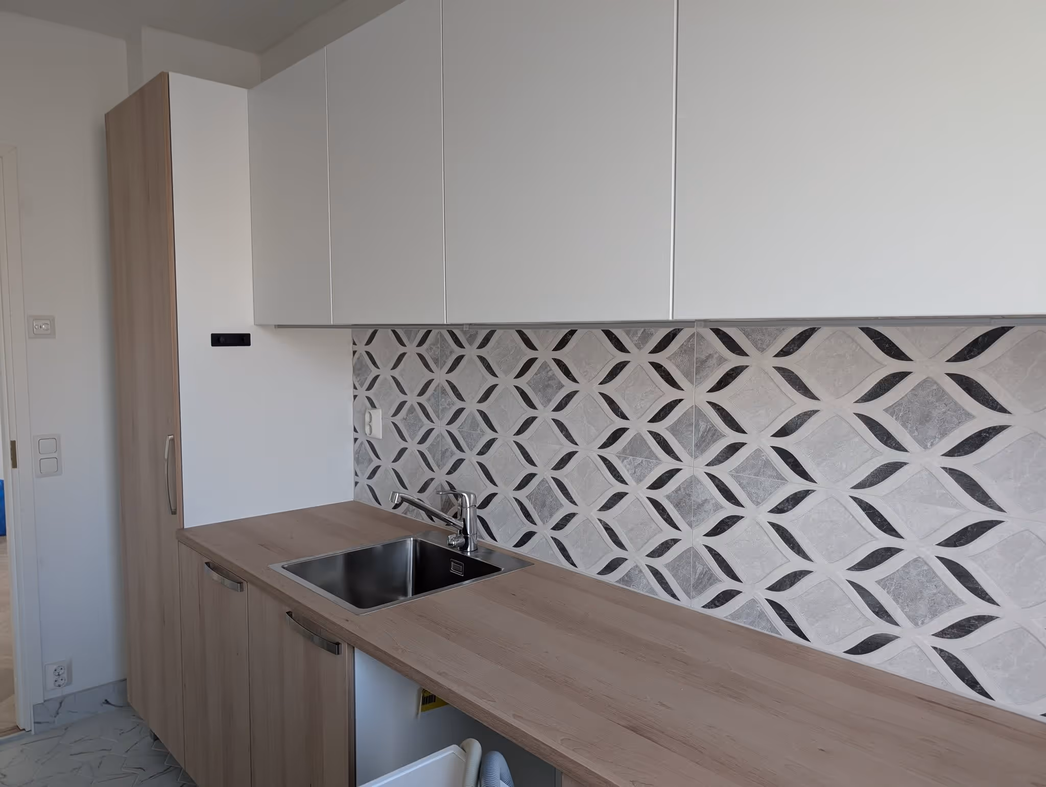 Moderni keittiö, jossa on vaaleat puiset työtasot, valkoiset yläkaapit ja koristeellinen harmaan ja mustan kuvioitu laattaseinä.