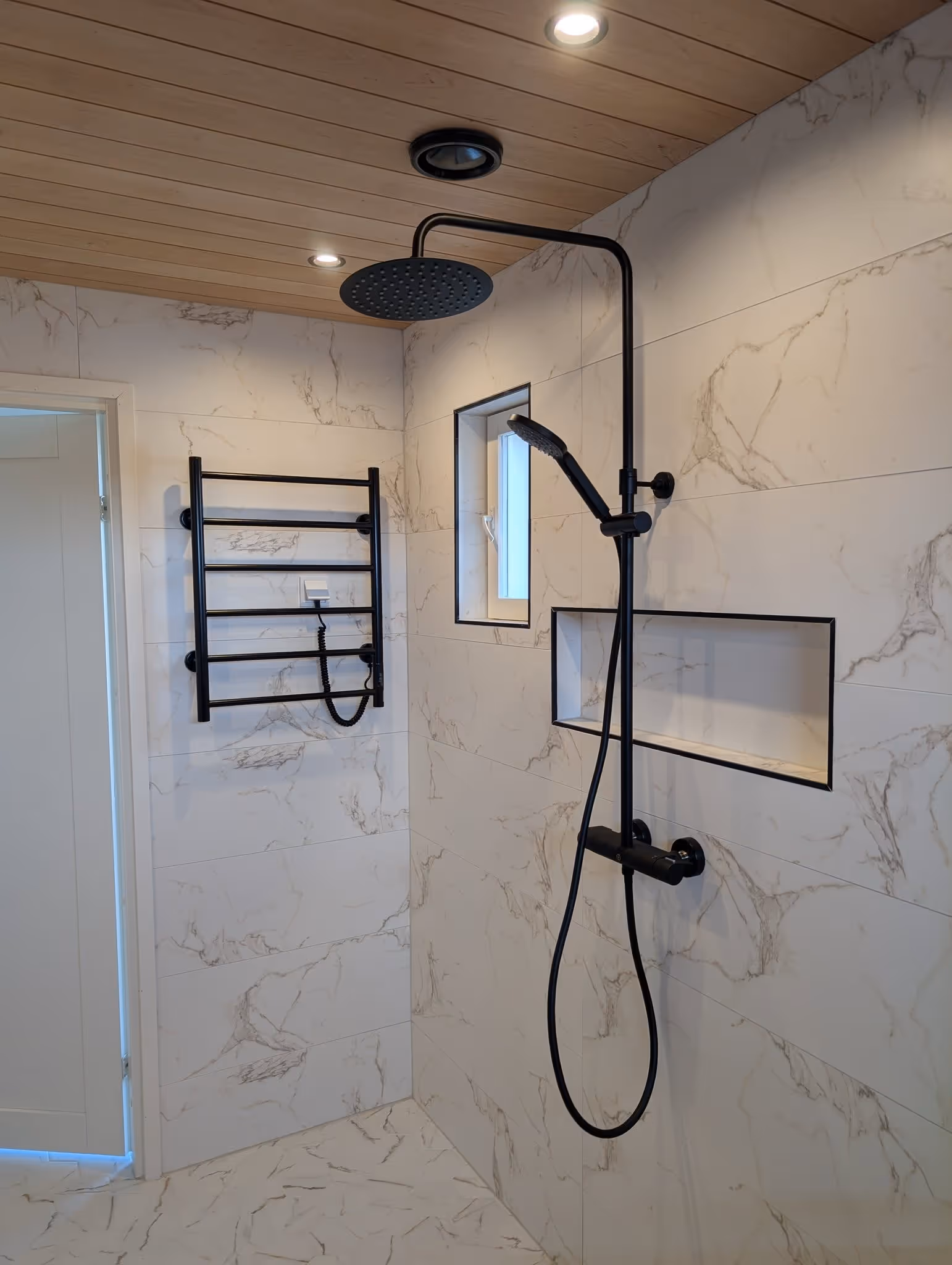 Moderni kylpyhuone, jossa musta sateenvarjopääsuihku ja käsisuihku valkoisissa marmorialaatoissa, katossa puupaneelit ja musta pyyhekuivain seinällä.