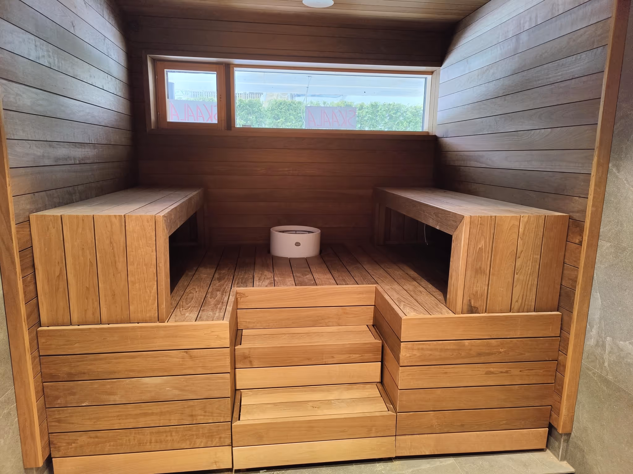 Puinen sauna, jossa kaksi penkkiä vastakkain ja ikkuna, josta näkyy ulkona kasvavia pensaita.