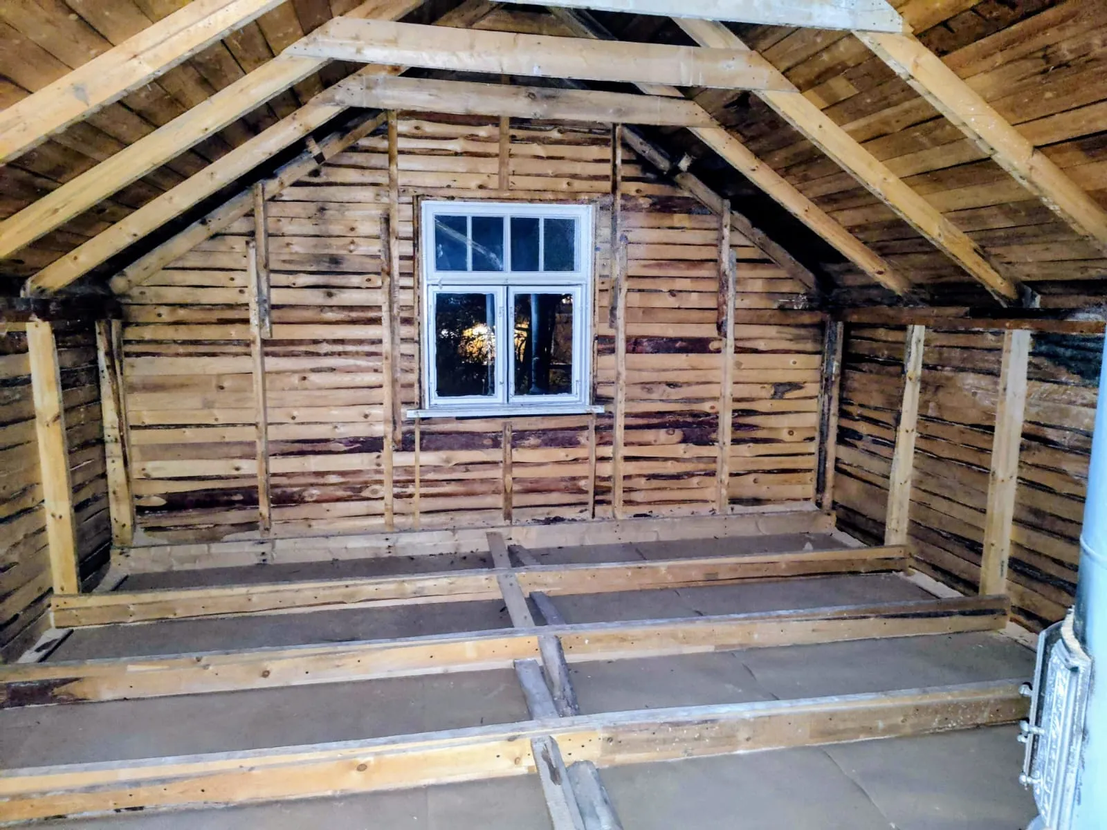 Puun runkojen raamit ullakkohuoneessa ikkunan alla ja vaaleat kattoparrut.