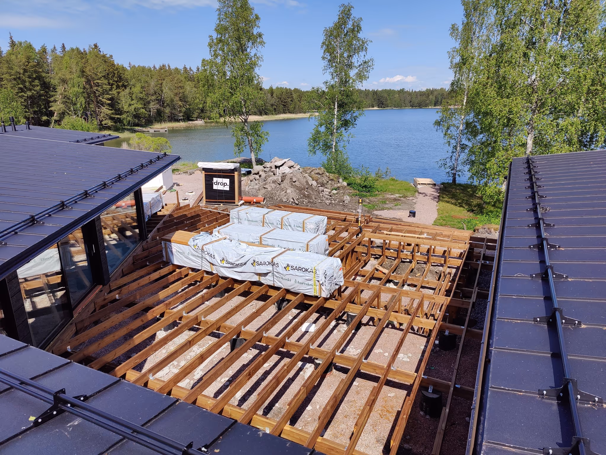 Rakenteilla oleva puulattia kahden talon välissä järvinäköalalla ja metsää taustalla.