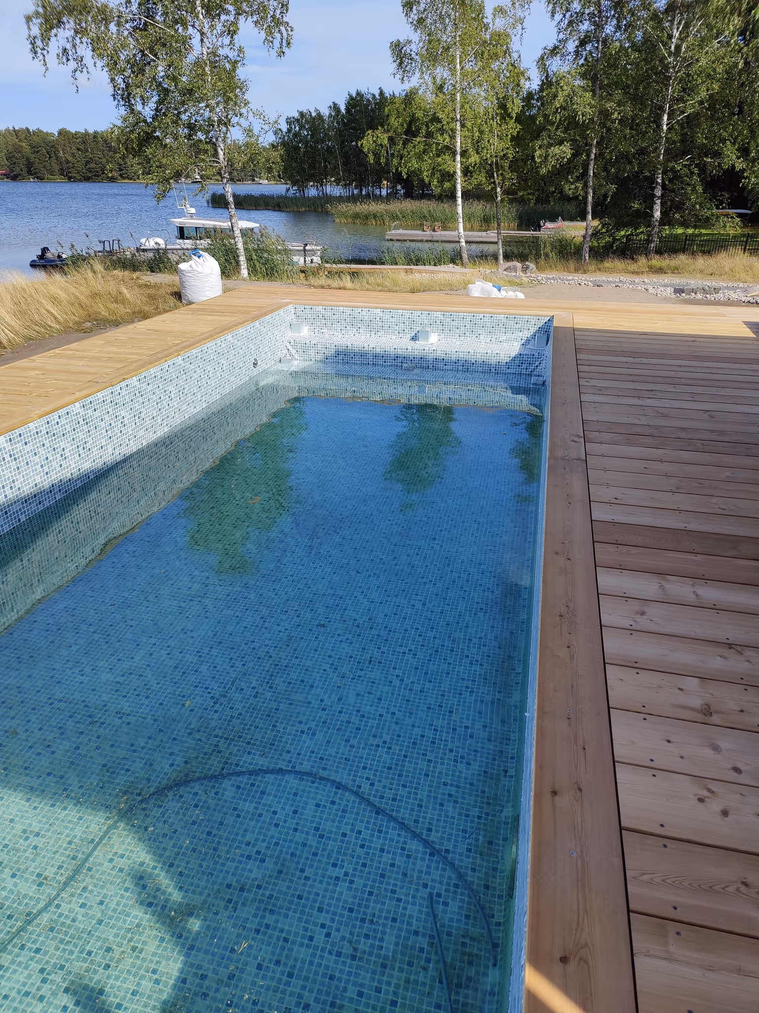 Uima-allas sinisillä mosaiikkilaatoilla ja puulaituri järven rannalla, ympärillä puita.
