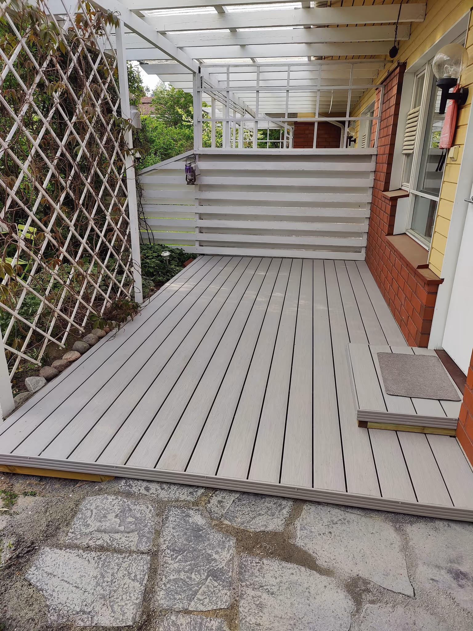 Vaalea puupaneelilattia, jossa pieni askelma ja harmaa matto talon punatiilisen seinän vieressä ja valkoinen ritiläkasvirakenne vasemmalla.