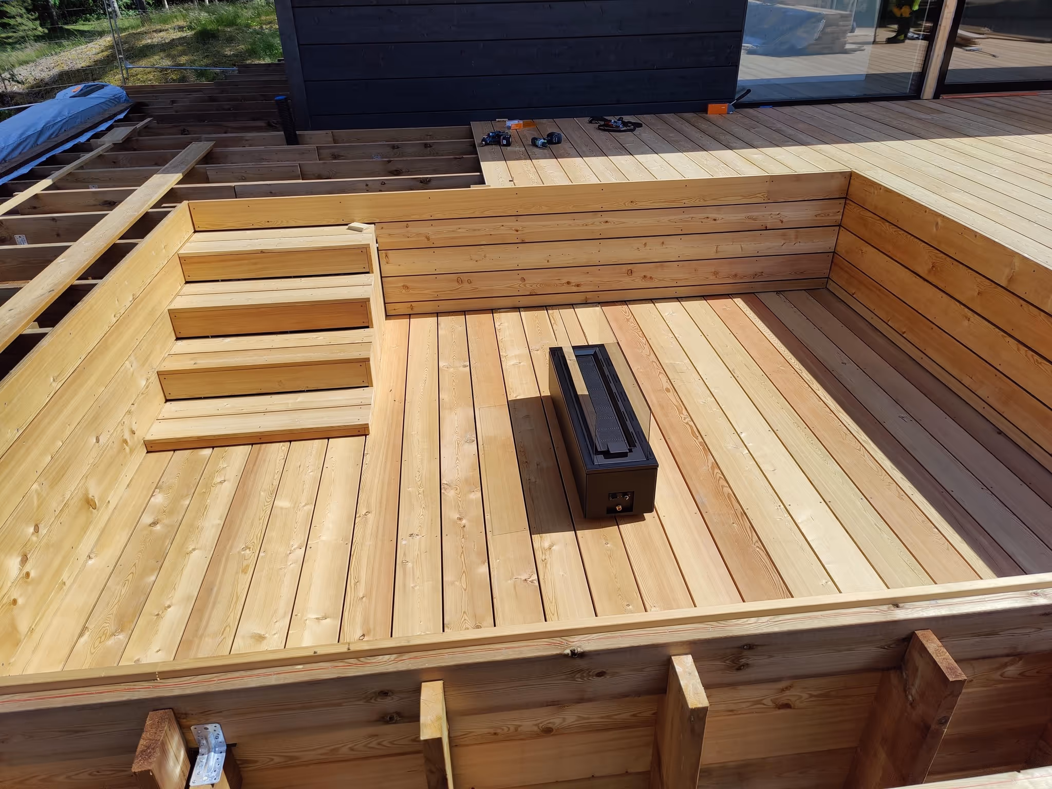 Ulkona puisesti rakennetun kuopan muotoinen terassin osan askelmat ja keskellä musta suorakulmainen laite.