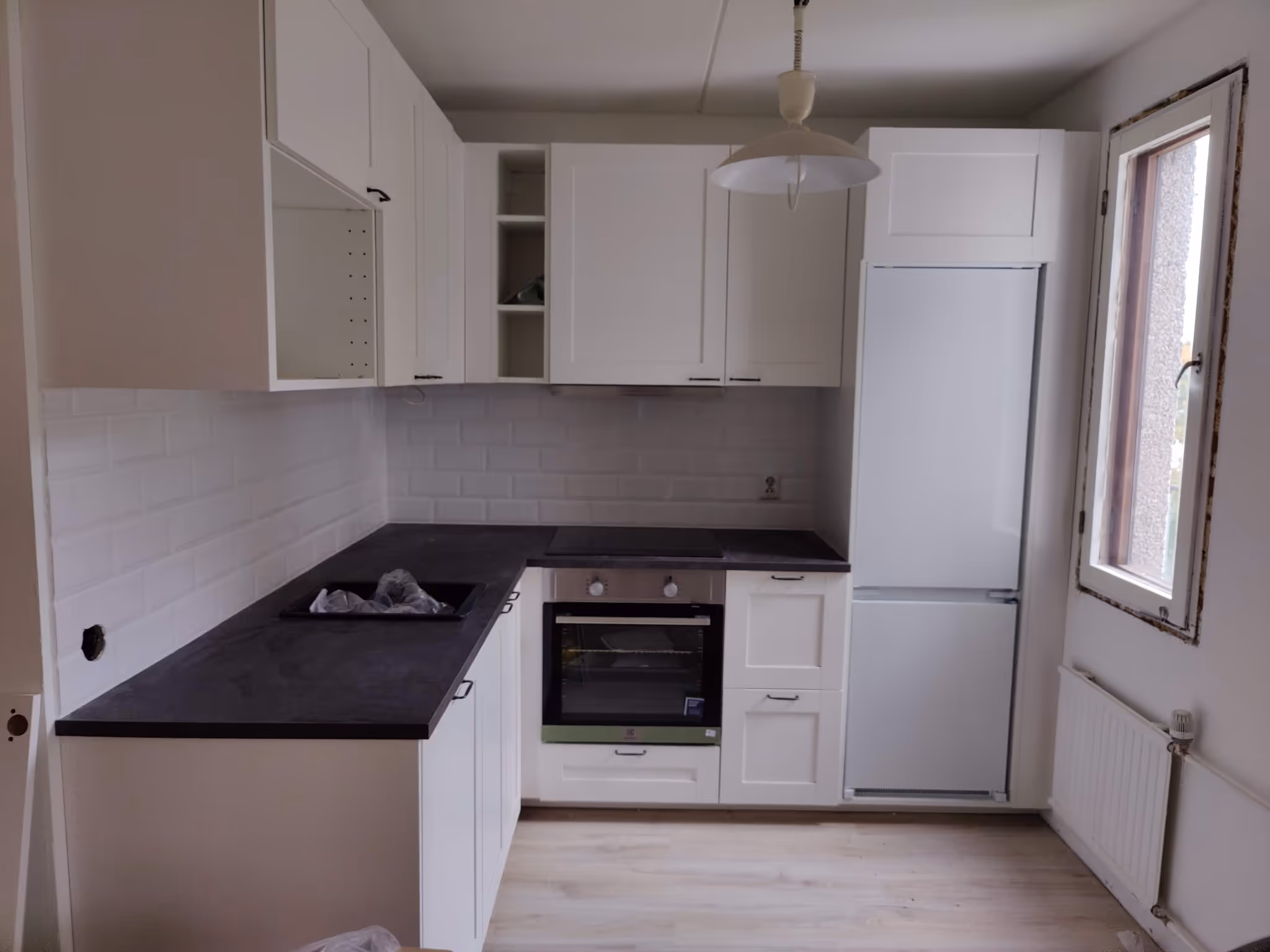 Pieni moderni keittiö, jossa valkoiset kaapit, mustat työtasot, uuni, liesi, jääkaappi ja iso ikkuna.