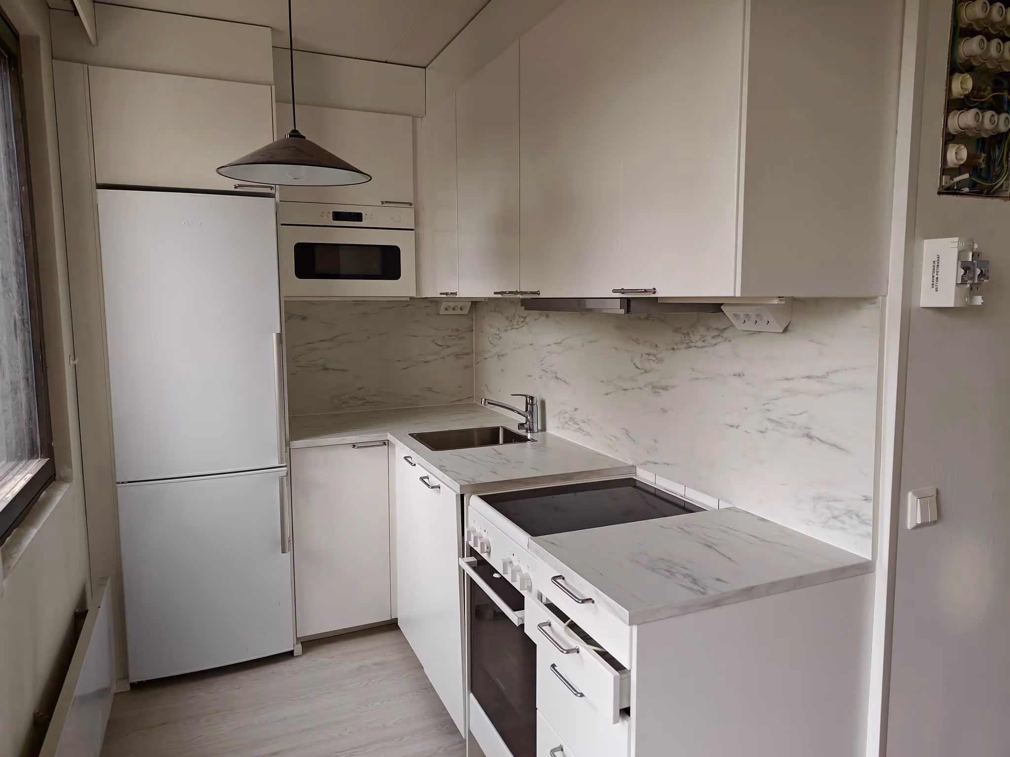 Moderni, pieni keittiö, jossa valkoiset kaapit, jääkaappi, uuni, mikroaaltouuni, allas ja marmorin näköinen työtaso ja välitila.