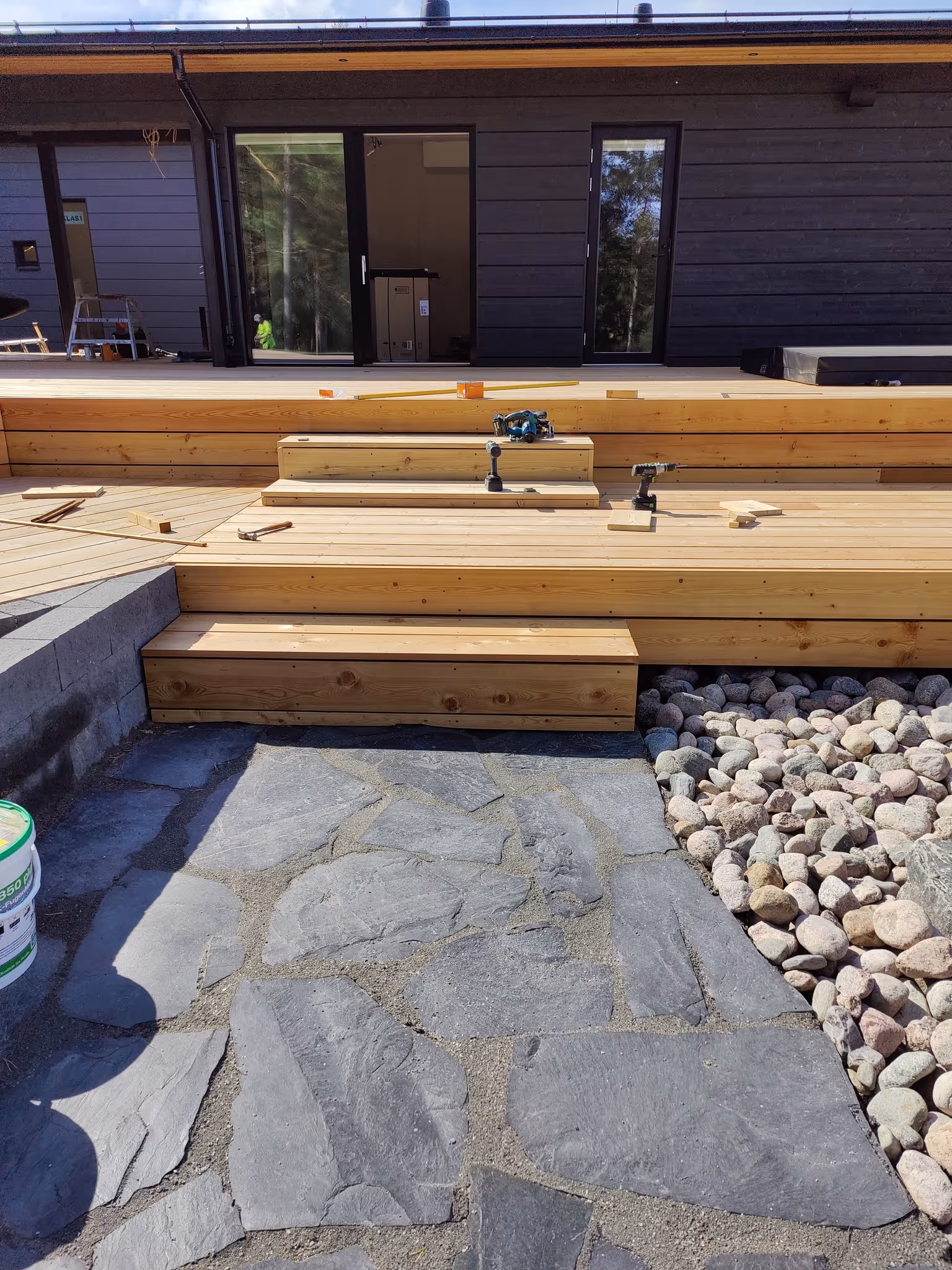 Ulkopuoli, jossa on keskeneräinen puinen terassi, lautoja, työkaluja, kivipolku ja kiviä.