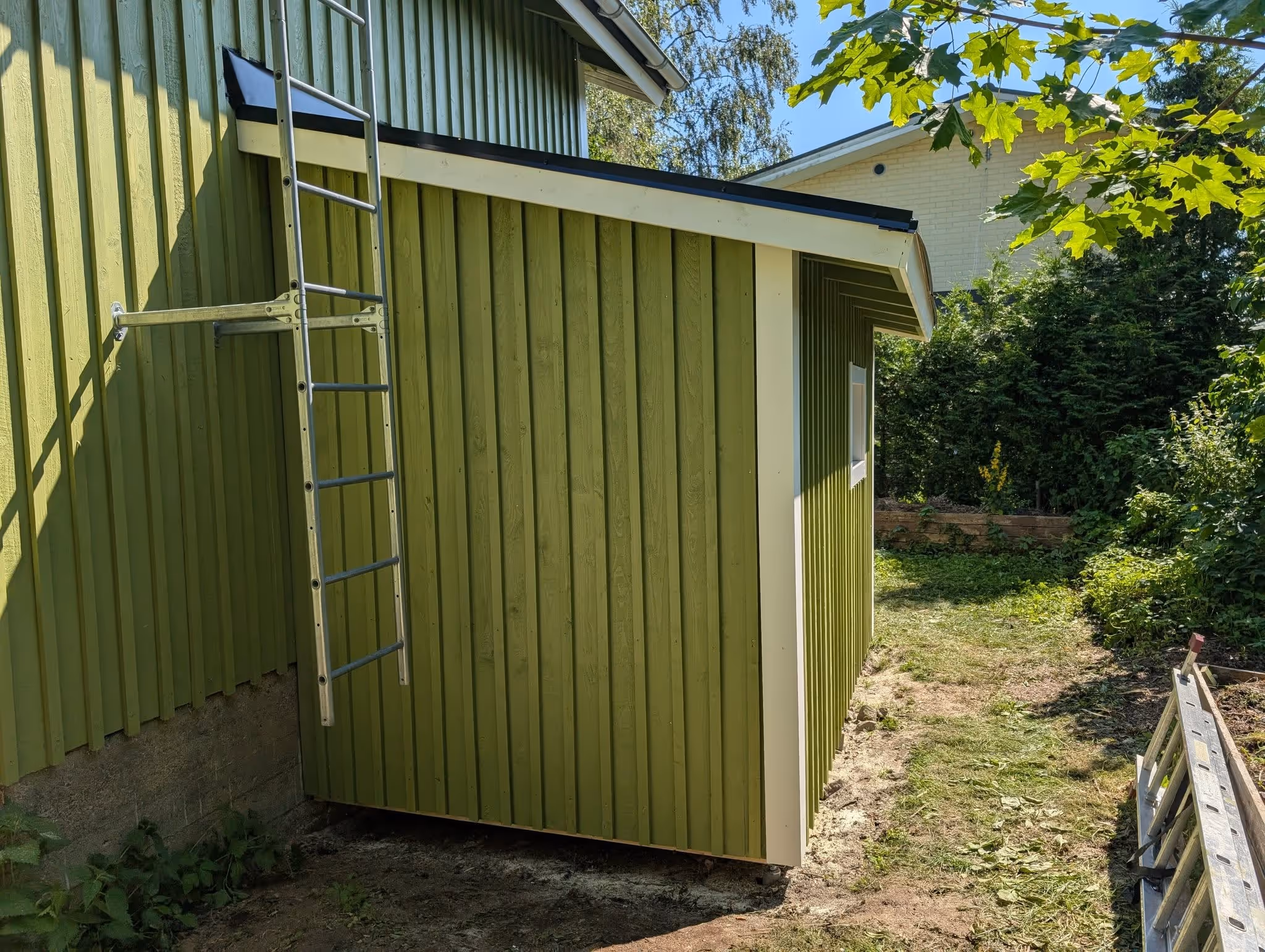 Vihreä puinen varasto tai aitta, jonka seinällä on kiinnitetty metallitikkaat, ympärillä nurmikkoa ja puita.